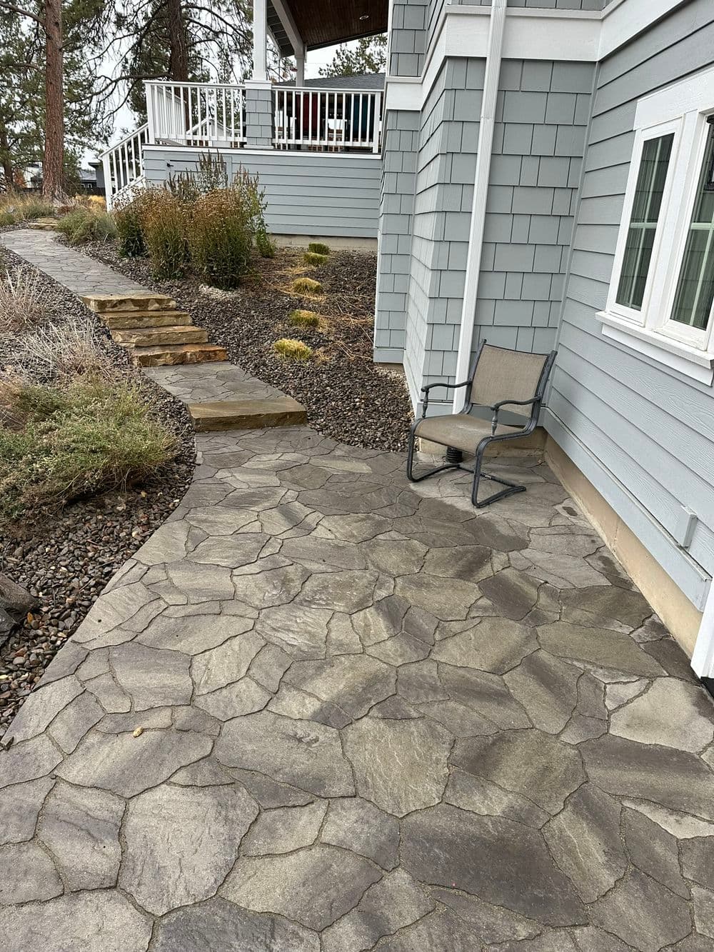 Irregular flagstone-pattern paver patio beside a gray shingled house, with sandstone slab steps climbing to a planted slope.