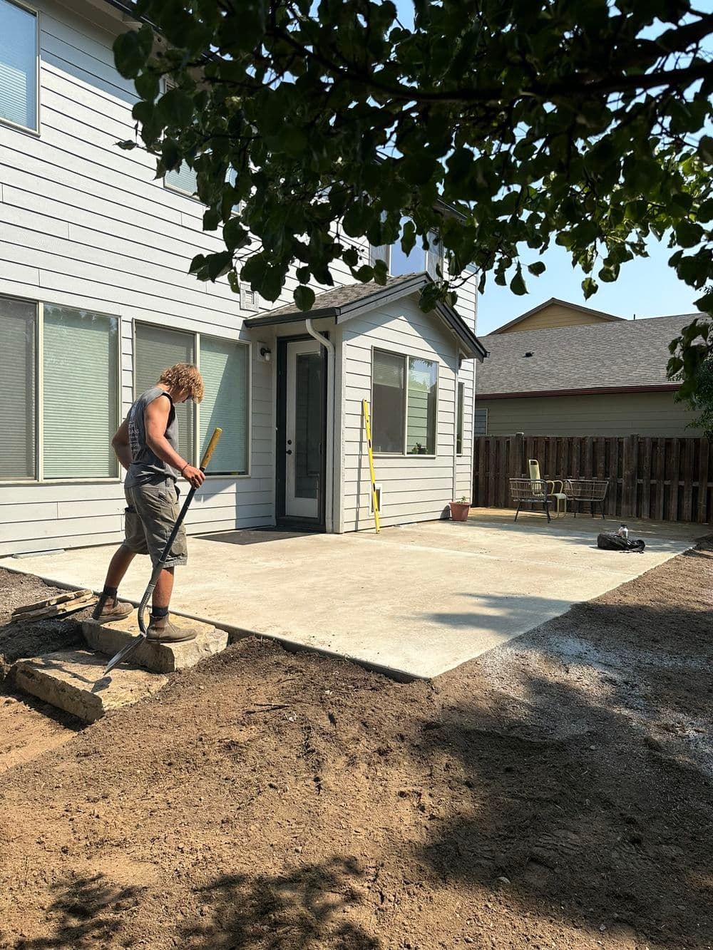 A crew member breaking up compacted ground with a mattock beside a concrete patio, graded soil ready for sod behind him.