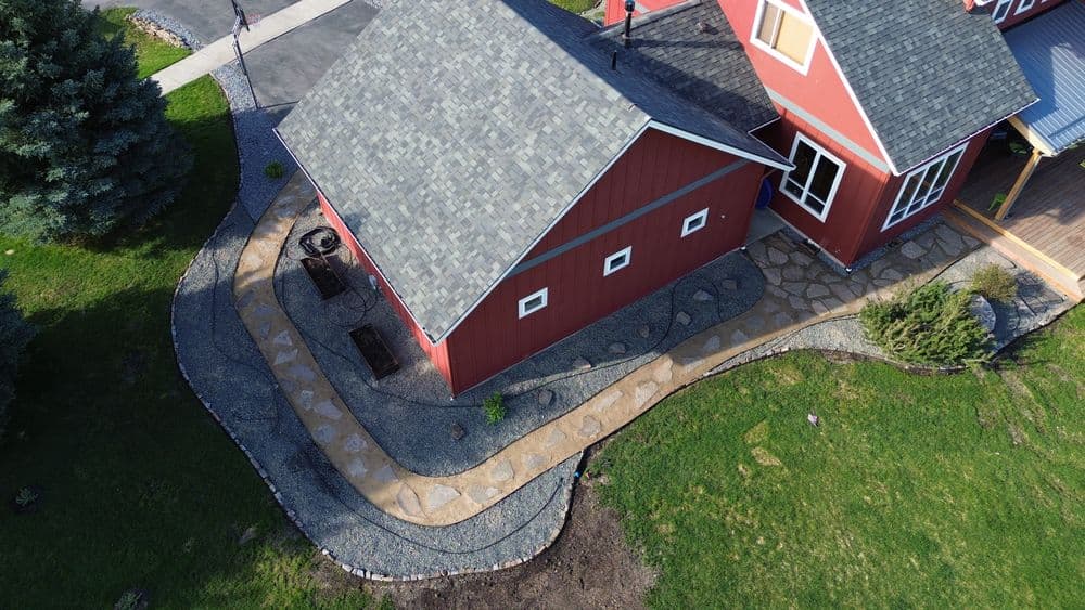 Overhead view of a curved decomposed granite path edged with boulders wrapping around a red outbuilding, with a flagstone landing at the house.