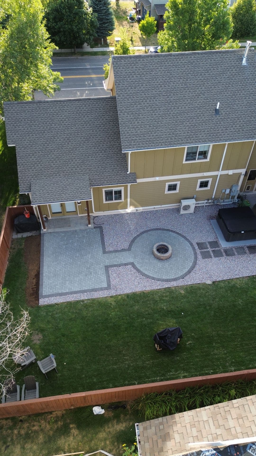 Overhead view of a backyard paver patio with a circular fire pit inset in a paver ring, gravel bands, a hot tub and new lawn.