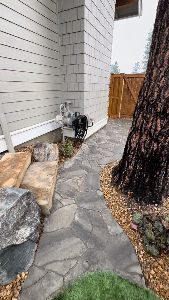 Flagstone-pattern paver walkway curving through a narrow side yard between a house and a fence, edged with gravel and boulders.