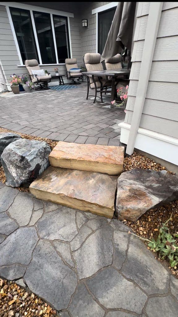 Two sandstone slab steps set between boulders, leading from a flagstone-pattern walkway up to a paver patio with furniture on it.
