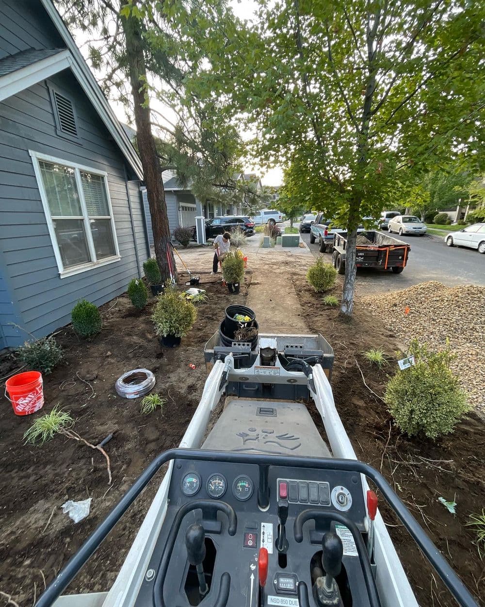 View from a landscaping machine showing a yard with trees, plants, and workers preparing the area.