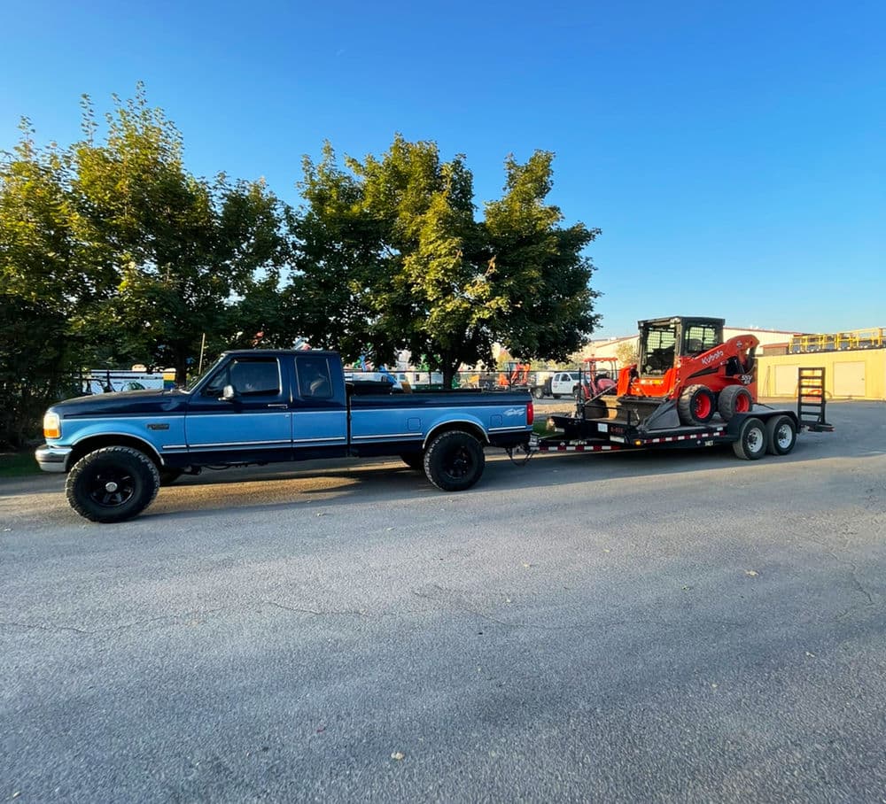 Blue pickup truck towing a trailer with construction equipment against a clear sky.