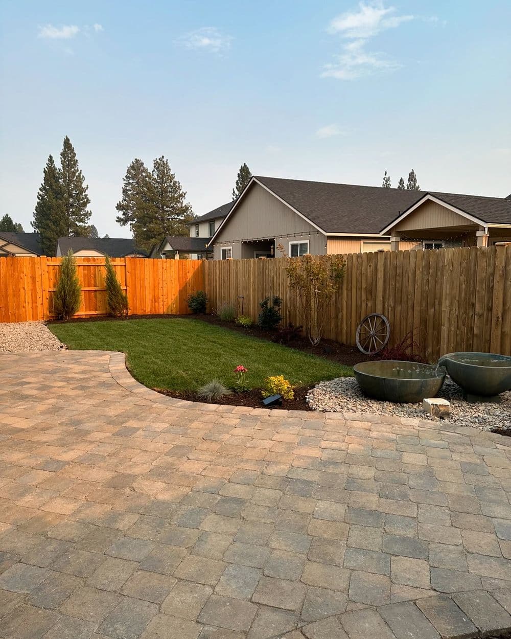 Backyard landscape with a fenced yard, green lawn, and stone patio, featuring decorative plants.
