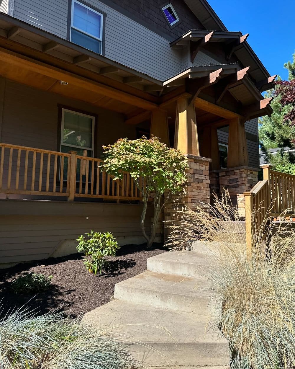 Front porch of a modern home with wooden railings, stone steps, and landscaped greenery.
