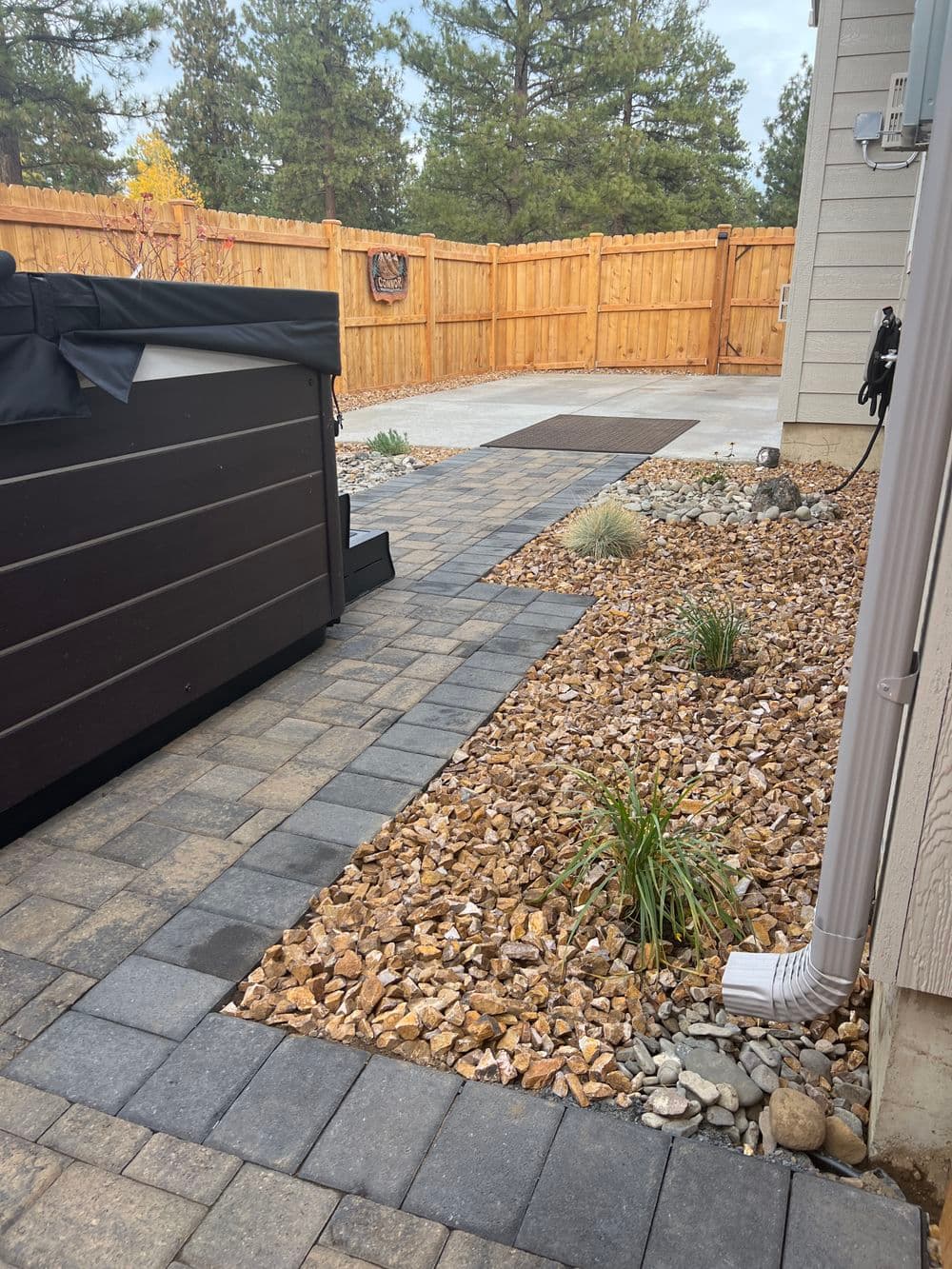 Paver walkway with a charcoal soldier-course border running past a hot tub to a concrete pad, with a crushed rock bed and ornamental grasses alongside.
