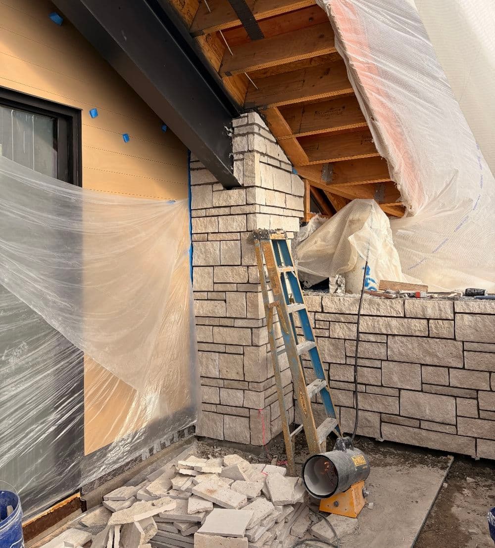 Cut limestone veneer going up on a column and low wall under a covered porch, with plastic sheeting and a heater running for cold-weather mortar work.