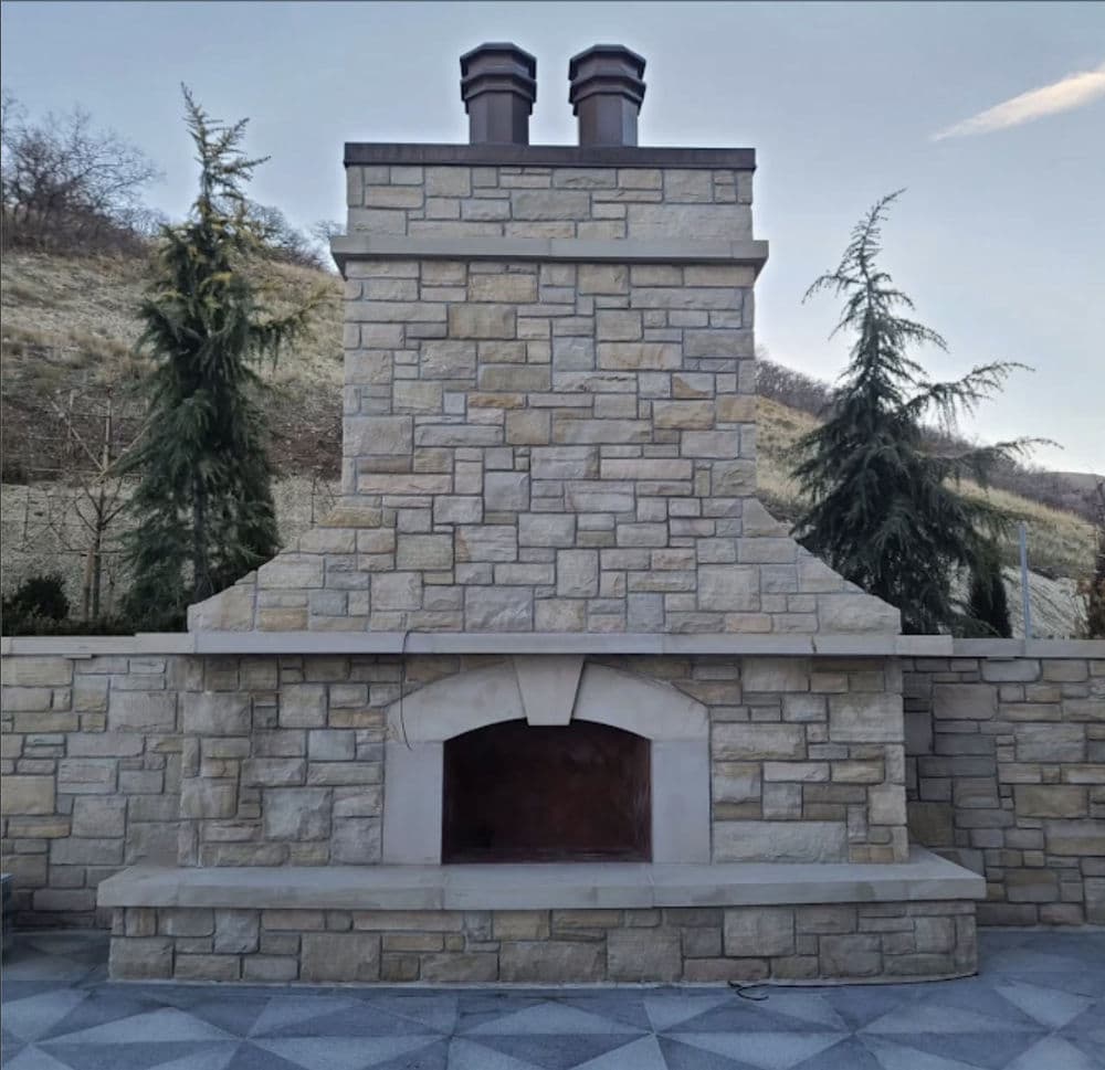 Outdoor stone fireplace with a tall chimney and two flue caps, built into a cut-stone wall with cast stone caps, on a paver patio below a hillside.