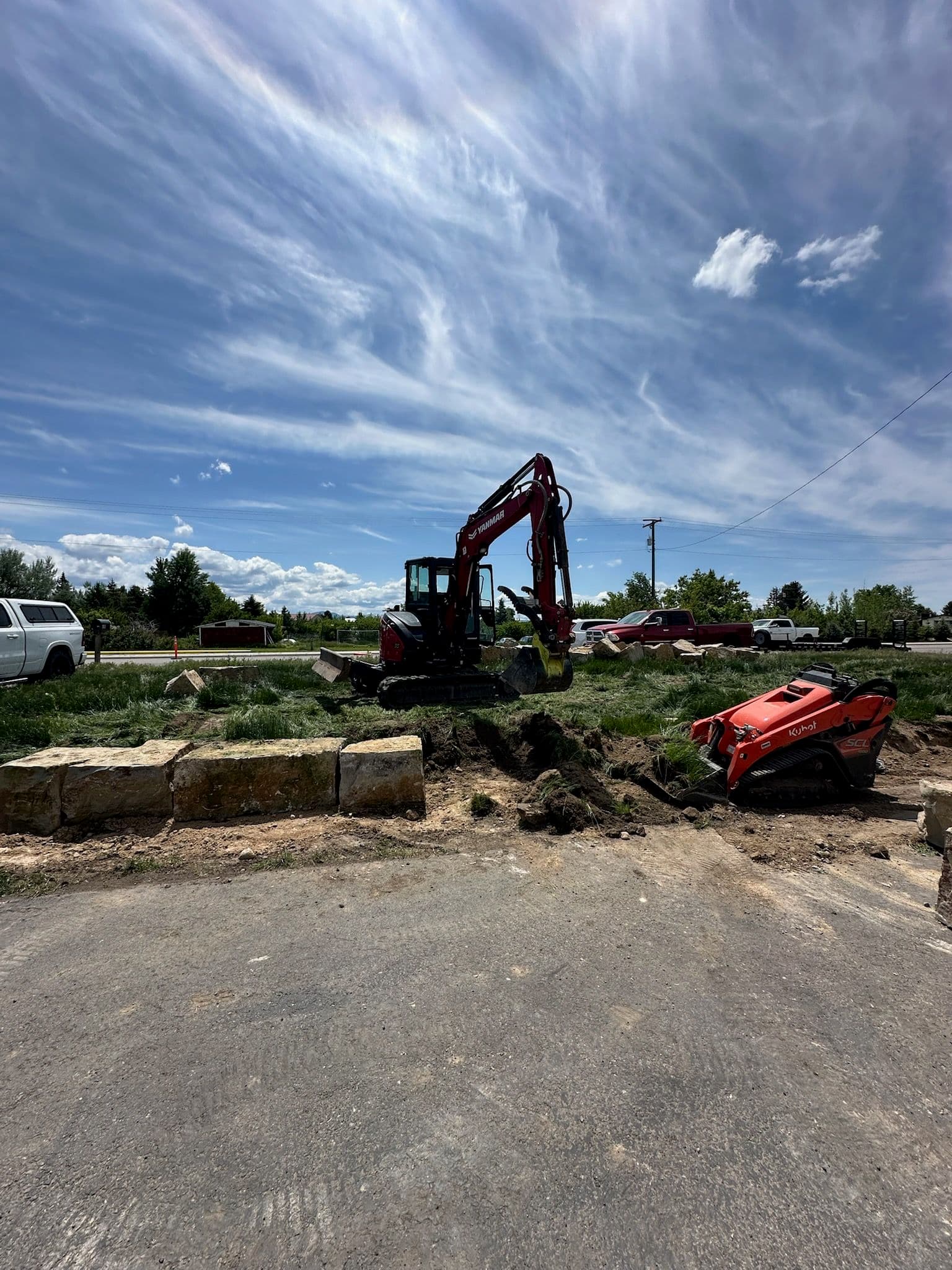 A course of quarried sandstone blocks being set as a retaining wall along a roadside, with an excavator and a compact track loader working behind it.