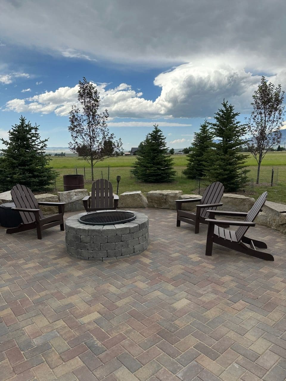 Herringbone paver patio with a round block fire pit, four Adirondack chairs and a boulder seat wall, open Montana field and mountains behind it.
