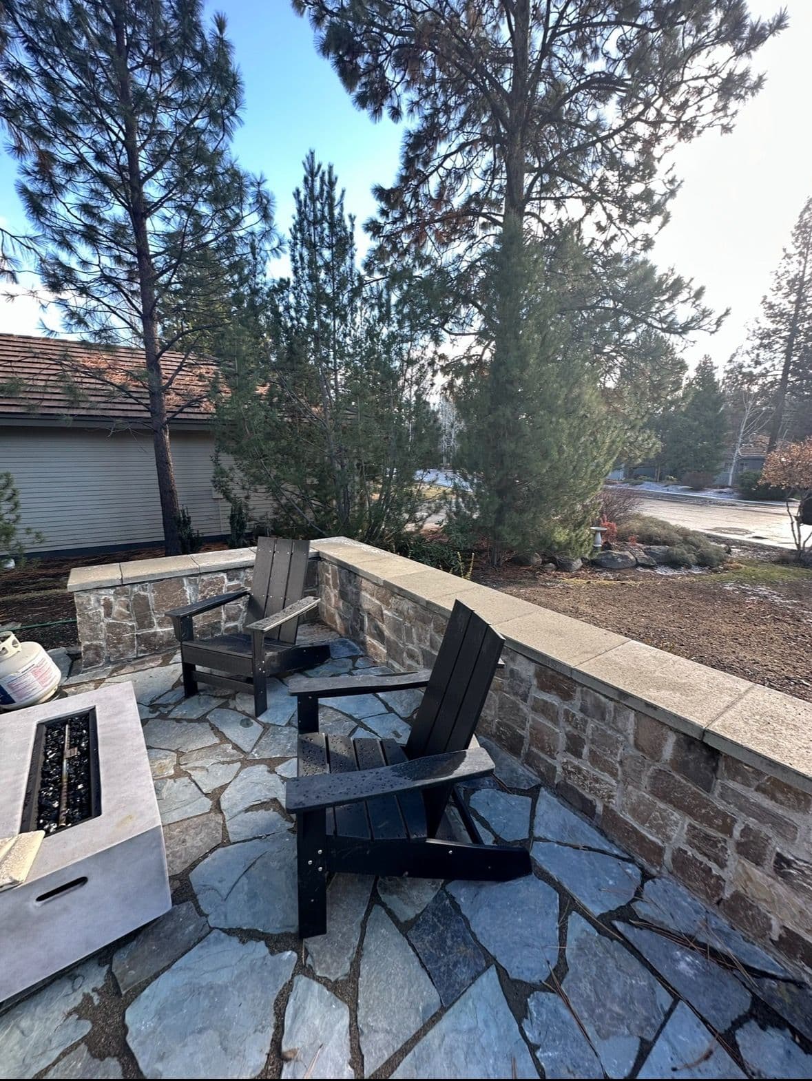 Flagstone patio with a mortared stone seat wall and flat cap stones, two chairs and a gas fire table, pines behind.